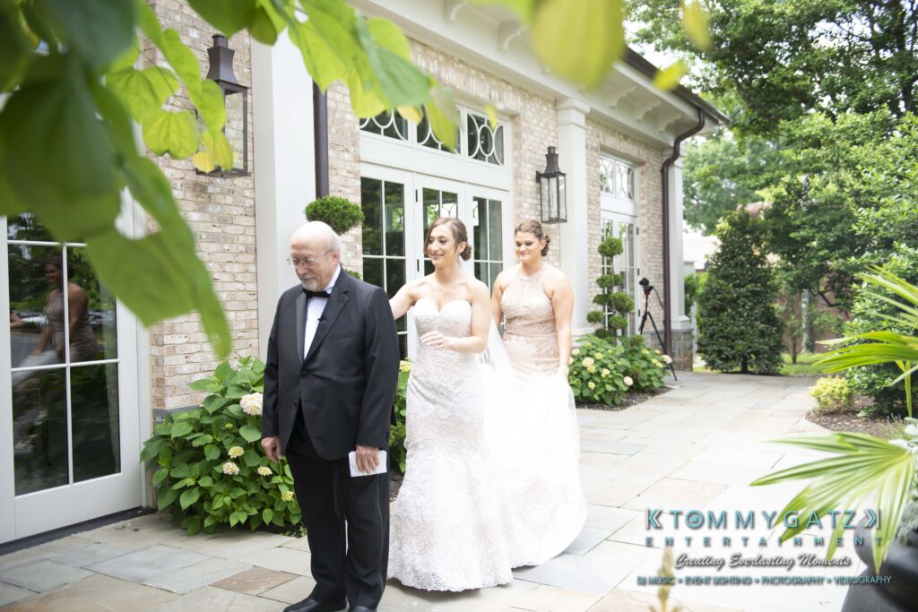 Bride in wedding gown holds hands with older man in a tuxedo outside a brick building. Two bridesmaids stand behind them on a stone path.