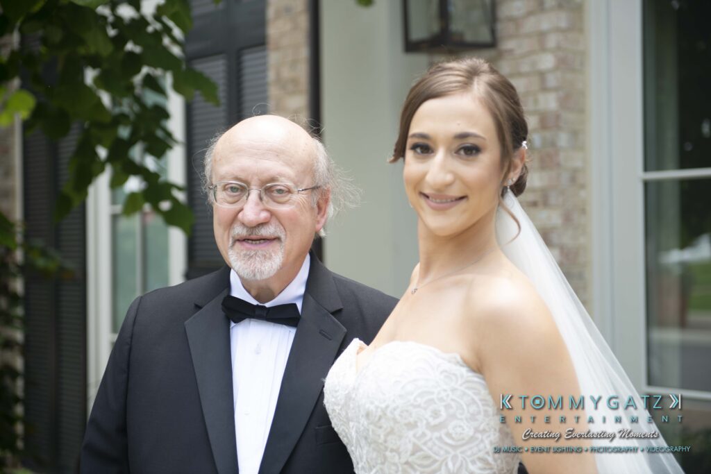 A bride in a white dress stands next to an older man in a tuxedo outside a brick building.