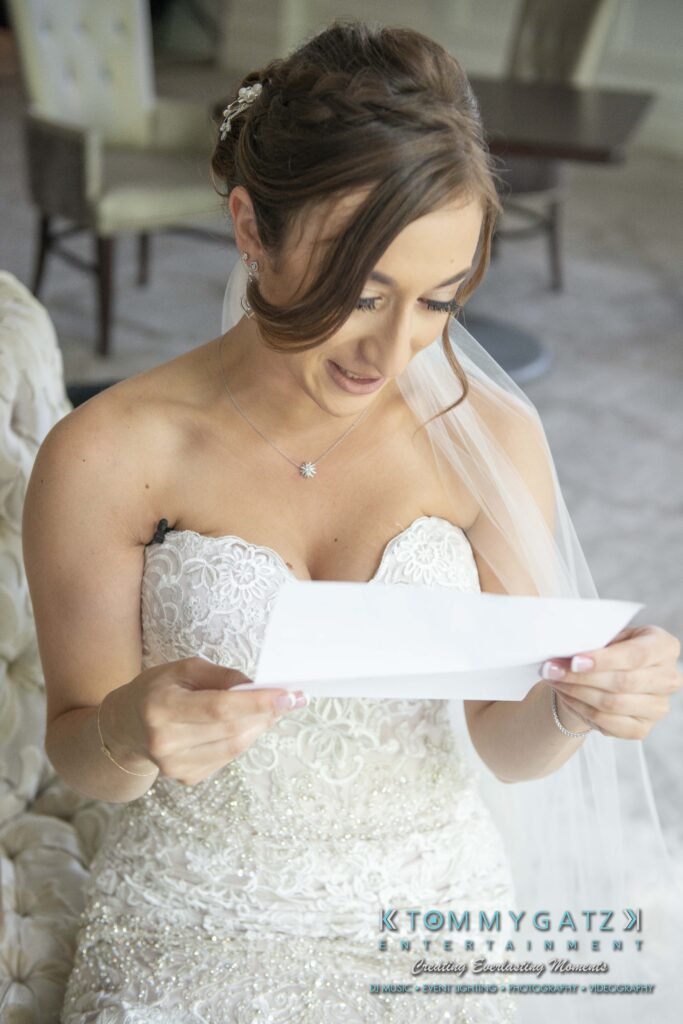 Bride in beaded white gown sits on a couch, reading a letter. Her hair is styled in a bun with a veil, and she wears a necklace. Logo at the bottom says "Tommy Gatz Entertainment.