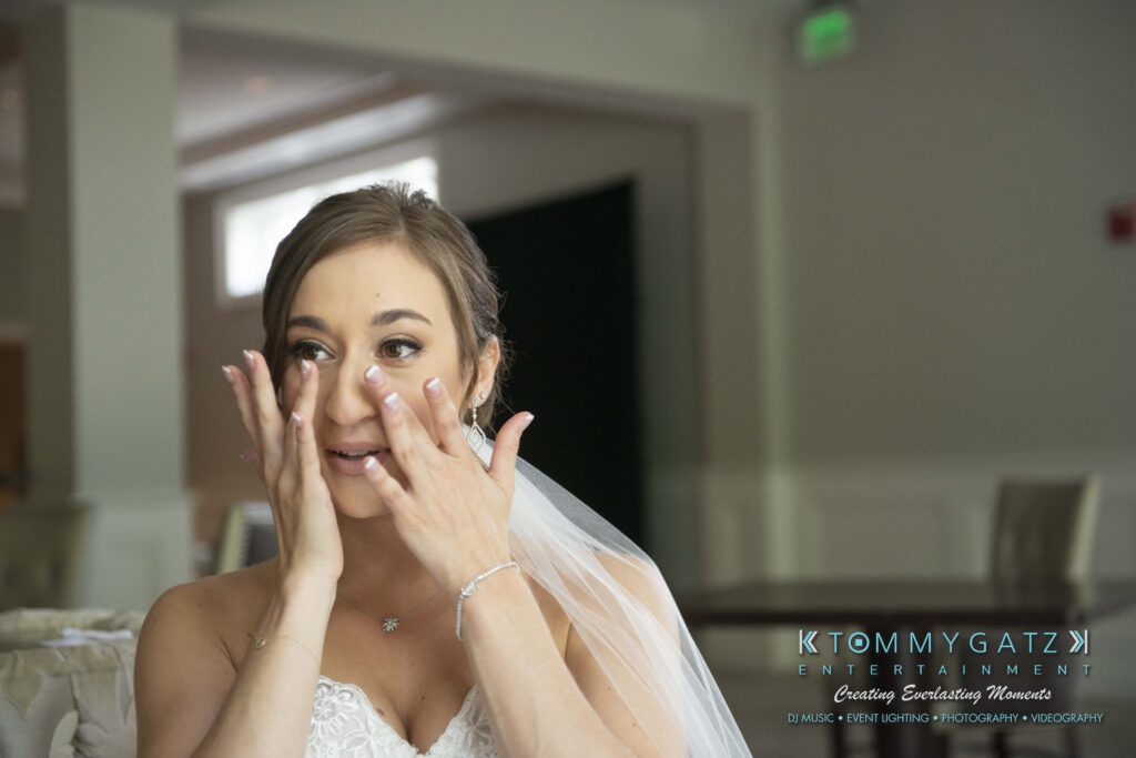 Bride in a wedding dress wipes tears from her eyes in an indoor setting, with faint logo in the bottom corner.