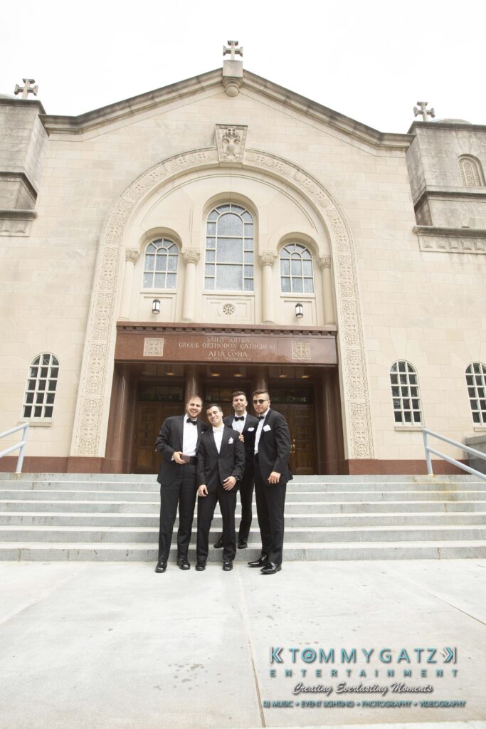 Four men in tuxedos stand on the steps of a large stone building with arched windows and a decorative facade.