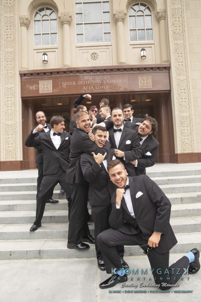 A group of men in tuxedos pose playfully on the steps of a building with arched windows and a Greek Orthodox Cathedral sign.
