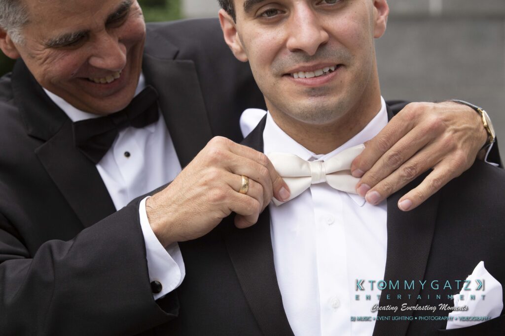 Urania-Aristidis-060218-PREP-Low-Res-WM-154 - TOMMY GATZ ENTERTAINMENT Two men in black suits share a moment. One adjusts the other's white bow tie, smiling. The logo "Tommy Gatz Entertainment" appears in the lower right corner.