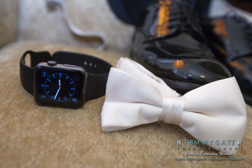 A black smartwatch, white bow tie, and shiny black shoes placed neatly on a cushioned surface.