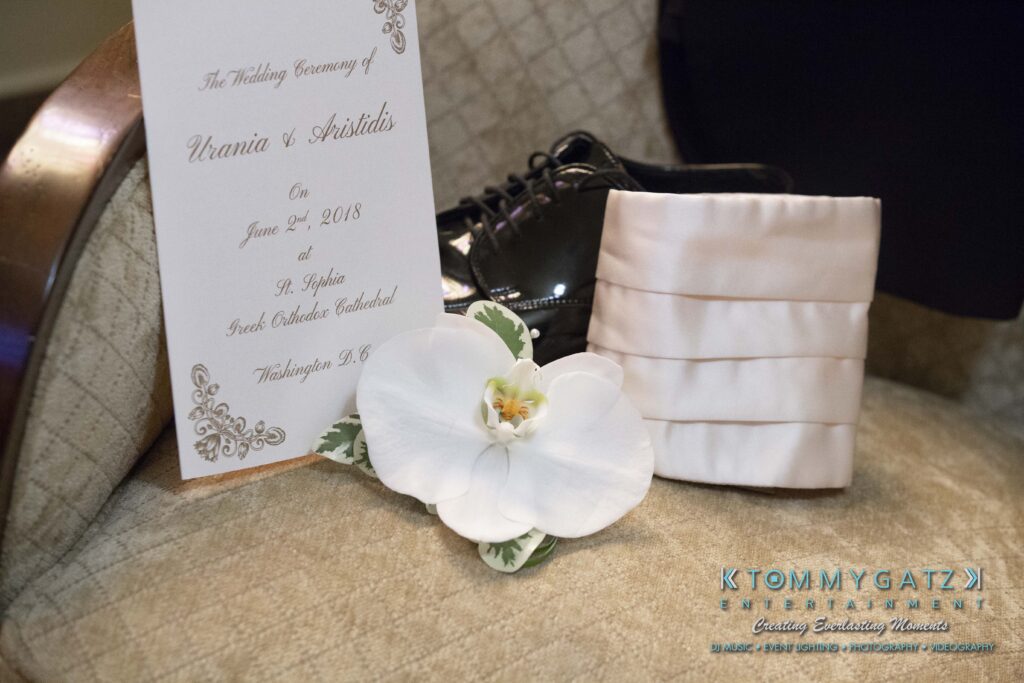 Wedding invitation card with a pair of black shoes, a white orchid, and a folded white sash on a beige chair.
