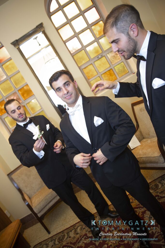 Three men in tuxedos inside a room. One adjusts his bow tie, another holds a flower, and the third touches the first man's shoulder. A window with a grid design is in the background.