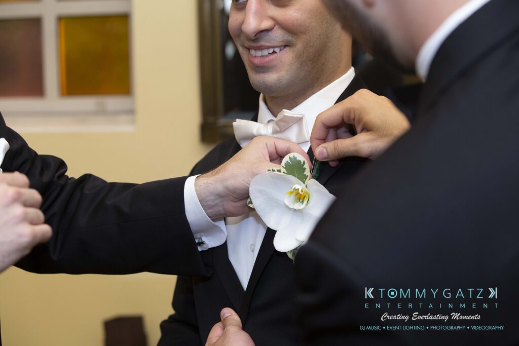 A man is having a white orchid boutonniere adjusted on his tuxedo by another person. The setting appears to be formal.