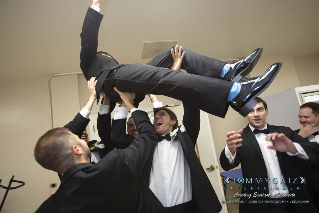 Group of men in suits joyfully lifting another man in an indoor setting.