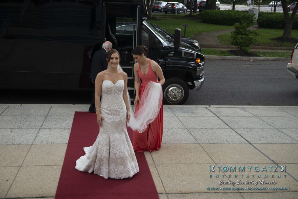Bride in a white gown walks on a red carpet toward a building entrance, followed by a bridesmaid in a coral dress holding the train of the dress, with a black shuttle bus in the background.