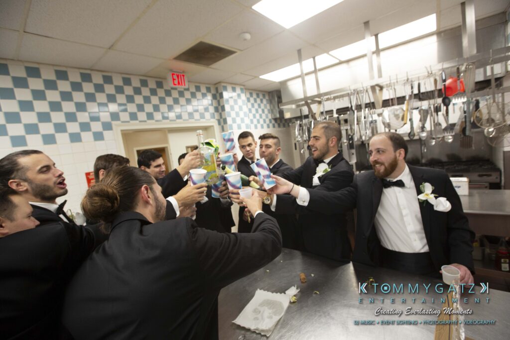 A group of men in tuxedos toast with drinks in a commercial kitchen.