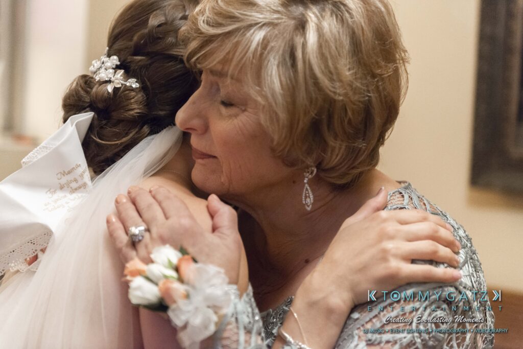 A bride and an older woman, both in formal attire, embrace warmly indoors.