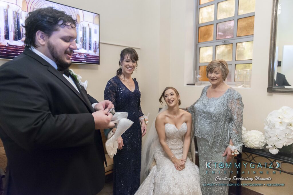 Four people in formal attire are in a room. A man in a suit appears to be speaking, while three women in gowns, one seated and wearing a wedding dress, listen nearby.
