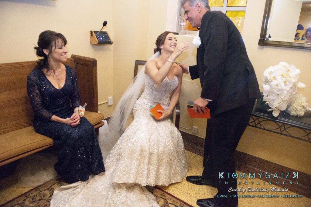 A bride in a white dress sits, holding an orange card, as a man in a suit leans down to kiss her cheek. A woman in a dark dress sits nearby, watching.