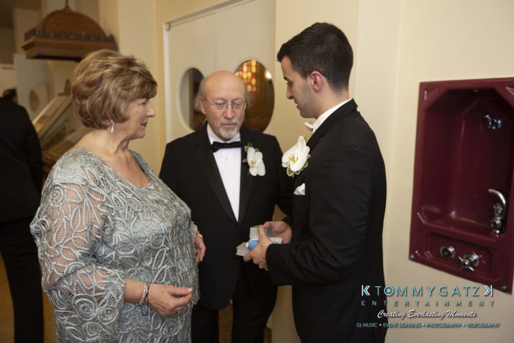 Three people in formal attire talking. One man, holding a small box, converses with a woman in a silver dress and an older man in a suit and bow tie.