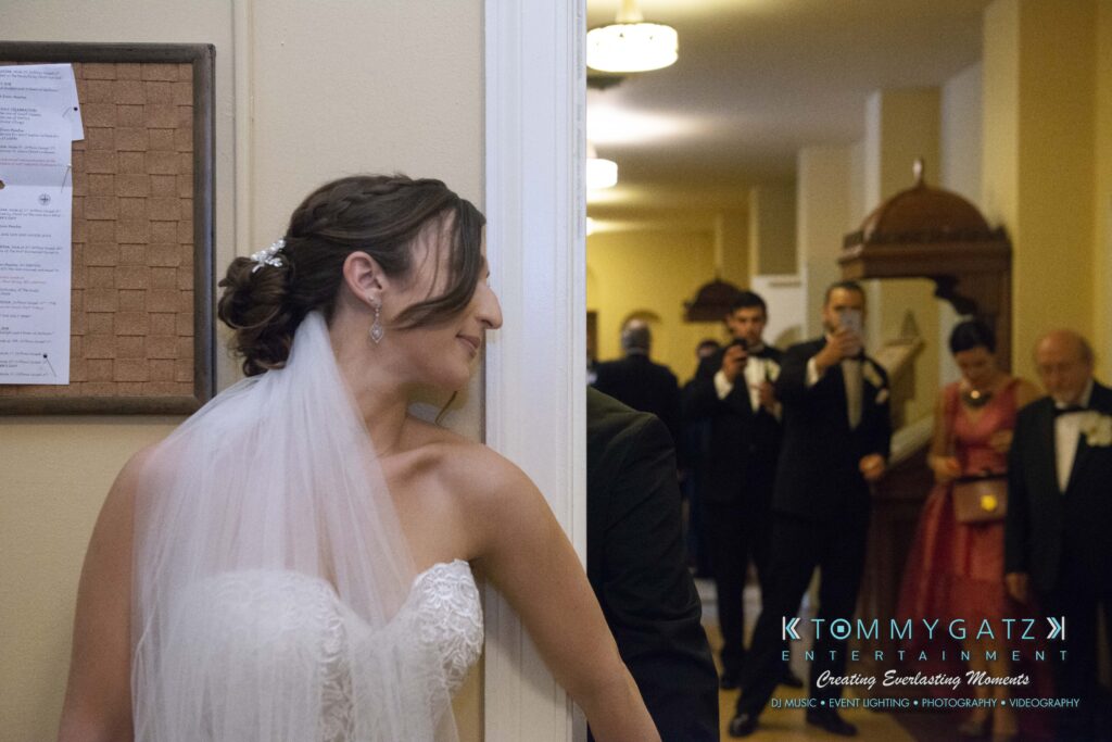 Bride in a white dress and veil stands near a doorway while a man is on the other side. People in formal attire are in the background, with some taking photos. A bulletin board is on the wall.