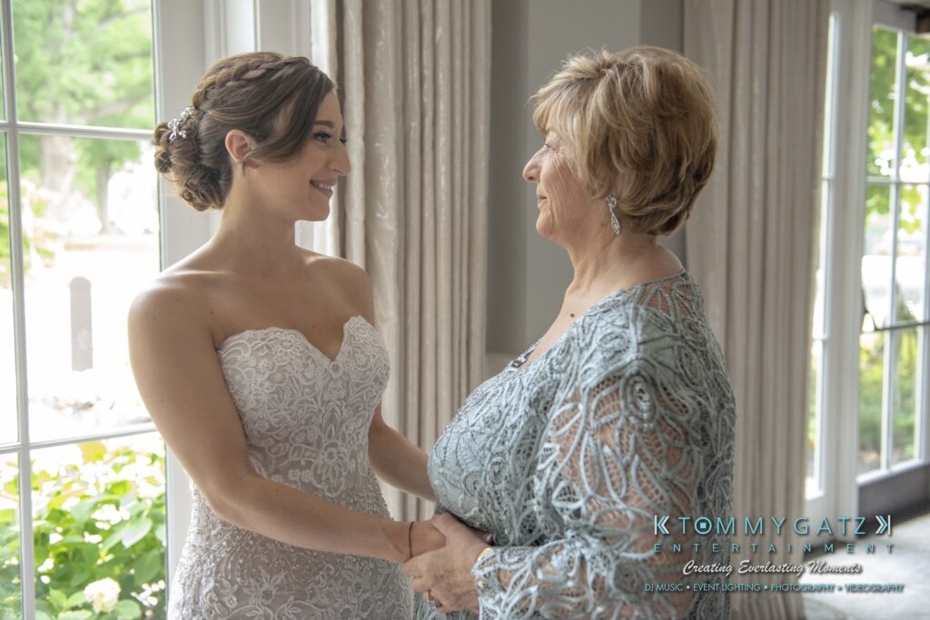A bride in a white lace dress holds hands with an older woman in a light blue dress while standing by a window.