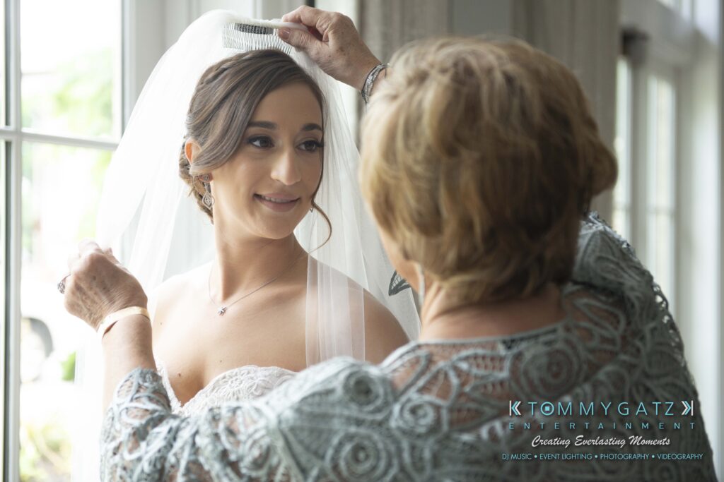 A woman adjusts a bride's veil in front of a window. The bride wears a white dress with lace details and earrings.