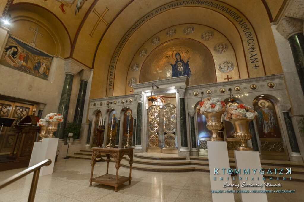 Interior of a church with a decorated altar, ornate gold doors, and a religious painting above. Floral arrangements and wooden chairs are present. Marble columns and arches are visible.