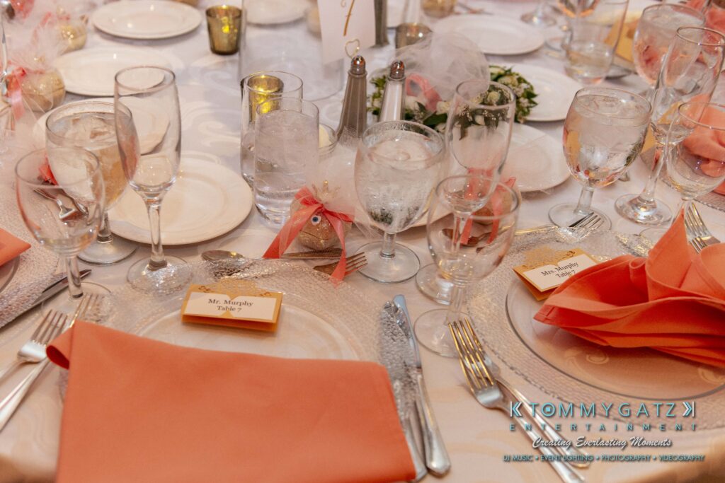 Elegant table setting with glassware, cutlery, and orange napkins. Name cards and decorative ribbons are present.