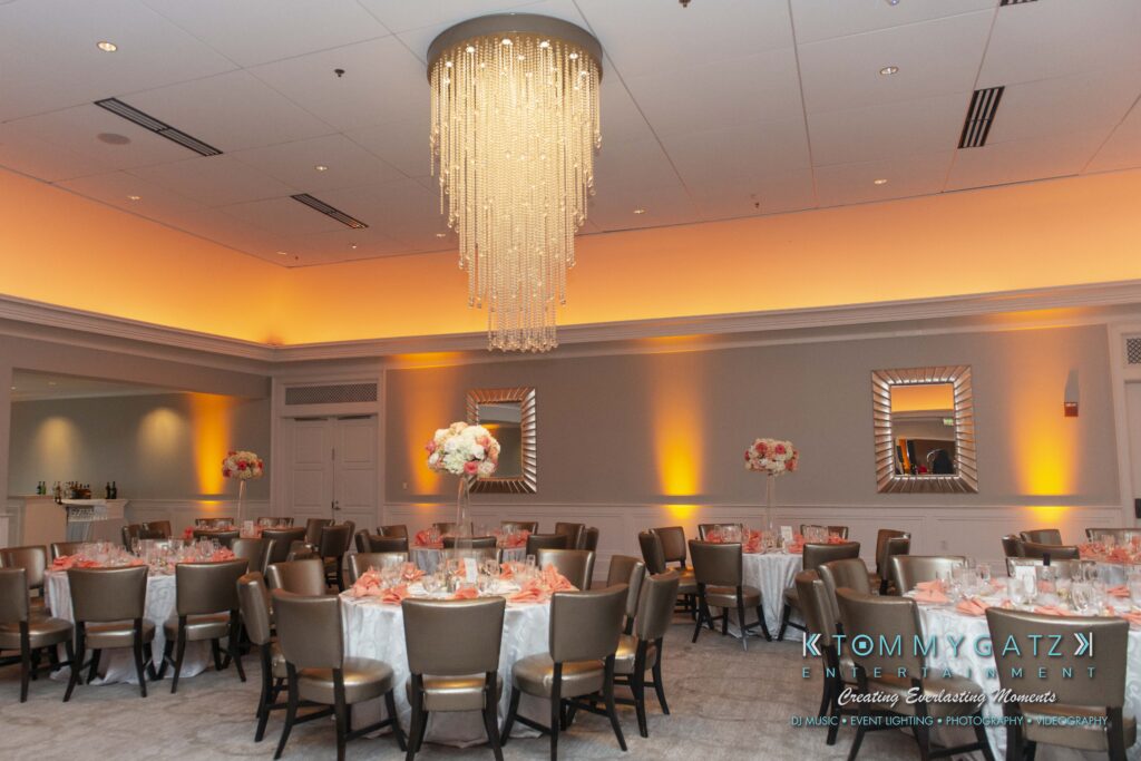 A banquet hall set for an event with round tables, beige chairs, peach table settings, floral centerpieces, mirrors, and a large chandelier.