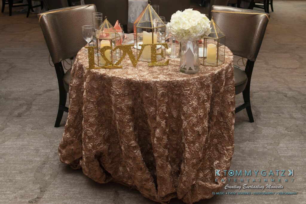 A round table with a brown floral tablecloth is set with glasses, white flowers, candles, and gold decorative elements, including one that spells "LOVE.