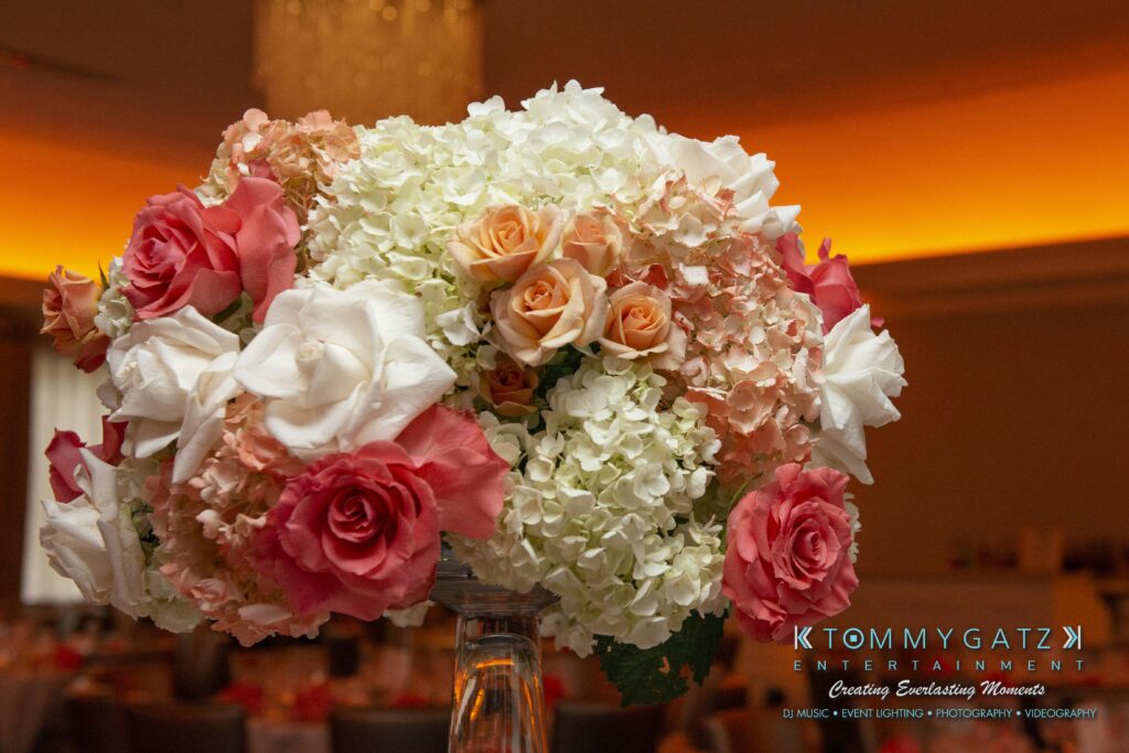 Floral arrangement with pink, white, and peach roses, white hydrangeas on a table. Room with an orange-lit ceiling in the background.