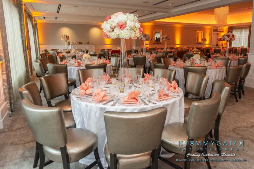 Urania-Aristidis-060218-RECEPTION-Low-Res-WM-630 - TOMMY GATZ ENTERTAINMENT Elegant wedding reception setup with round tables, white tablecloths, peach napkins, and floral centerpieces. Warm lighting enhances the sophisticated ambiance of the venue.