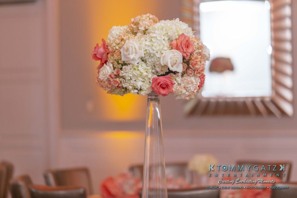 A tall vase holds a bouquet of white hydrangeas, pink and white roses. The background shows a blurred interior with warm lighting.