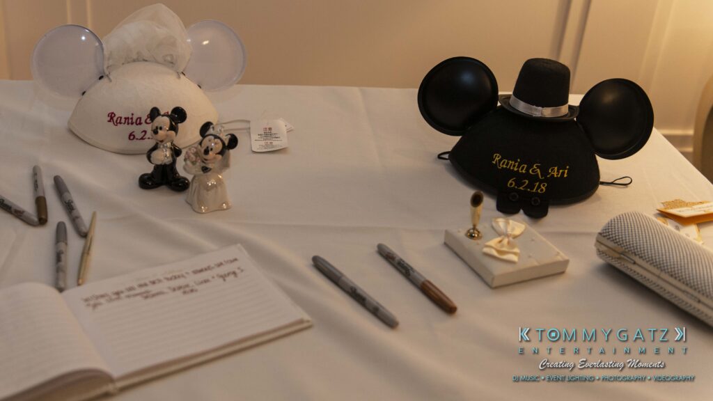 A table with two Mickey Mouse hats, a guest book, pens, miniature Mickey and Minnie figurines, and a tissue box, all arranged for a wedding-themed setup.