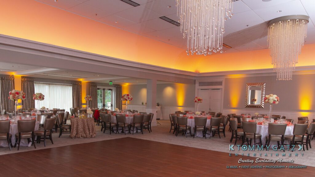 Spacious ballroom set for an event with round tables, floral centerpieces, a wooden dance floor, chandeliers, and warm overhead lighting.