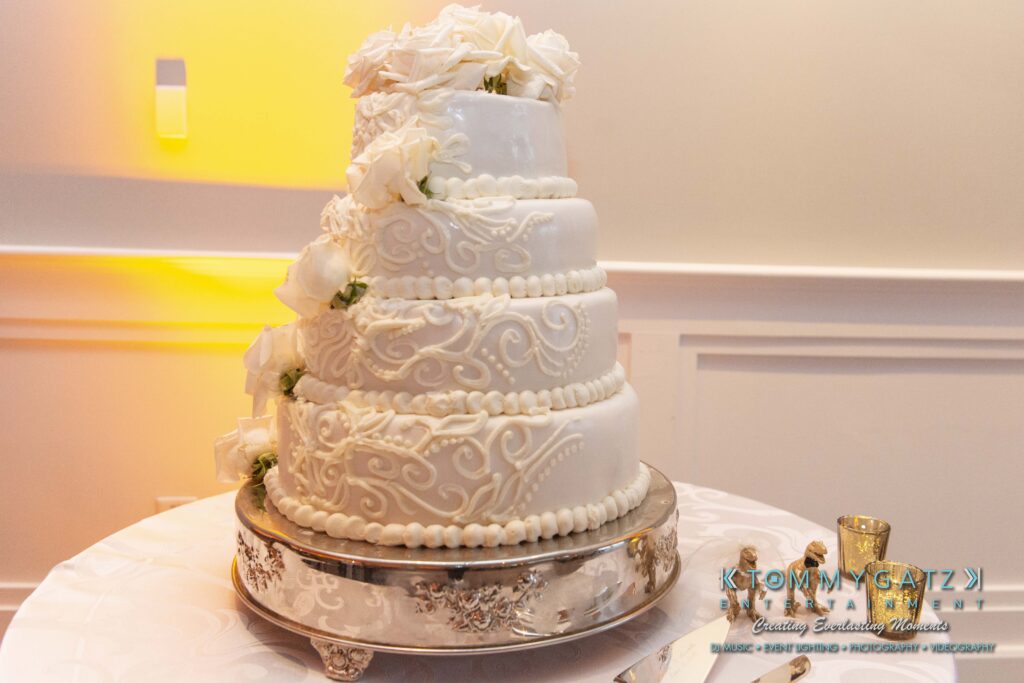 A five-tier white wedding cake with ornate swirl designs and white rose accents, displayed on a silver stand.