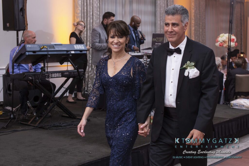 A couple in formal attire holds hands while walking. A band performs in the background, and guests are seated at tables.