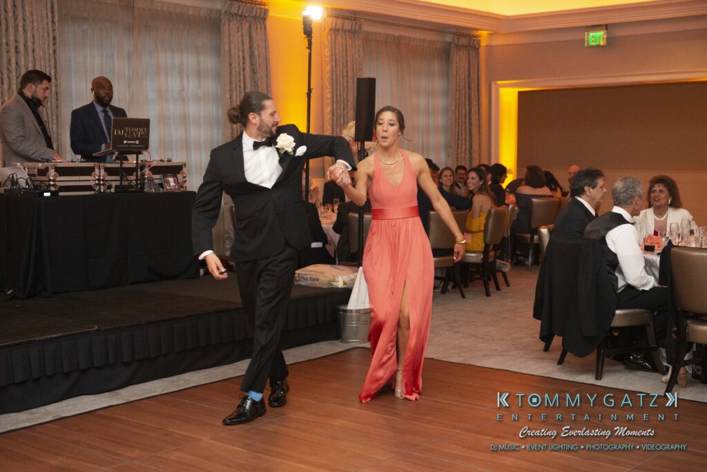 A man and woman elegantly dance on a ballroom floor, with a DJ and guests seated at tables in the background.