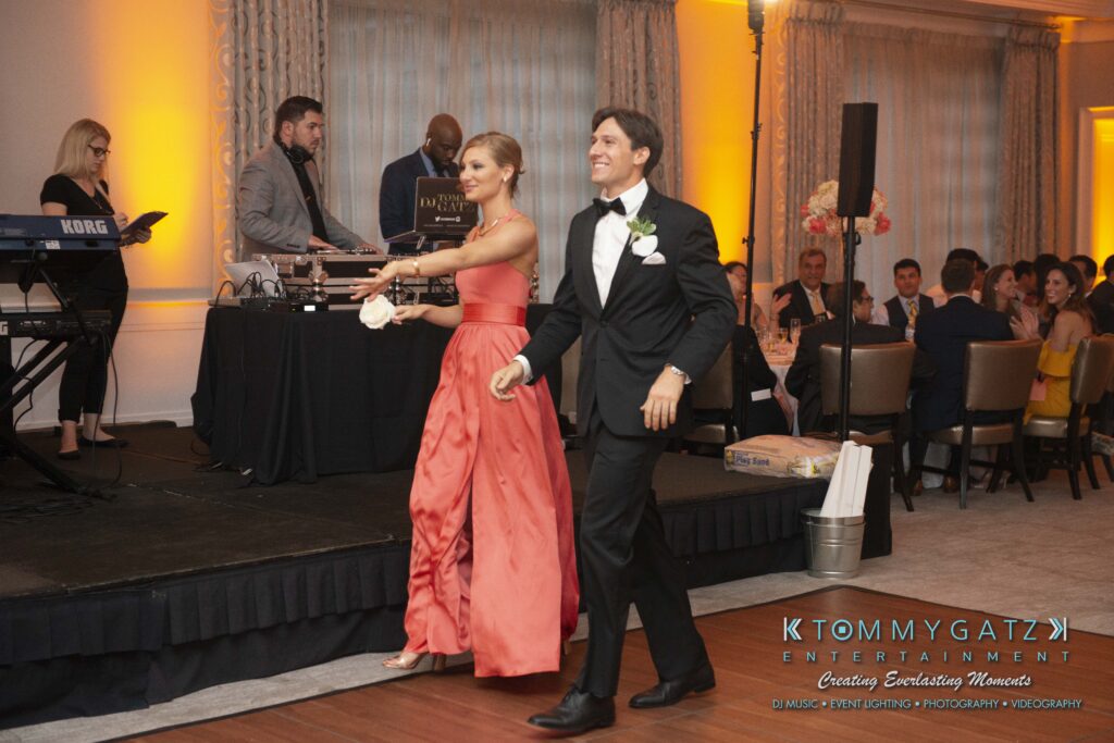 A couple in formal attire enters a venue, smiling. The woman wears a long pink dress, and the man is in a black tuxedo. Guests are seated at tables, and a DJ stands nearby.