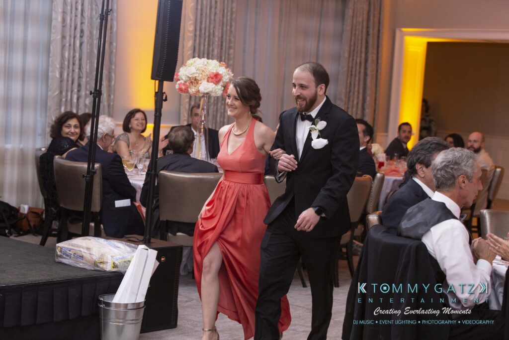 A woman in an orange dress and a man in a suit walk arm in arm at an indoor event with seated guests.