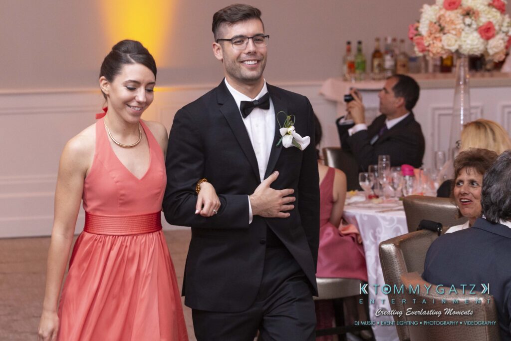 A man in a tuxedo and a woman in a coral dress walk arm in arm, smiling, at an indoor event with guests seated at tables in the background.