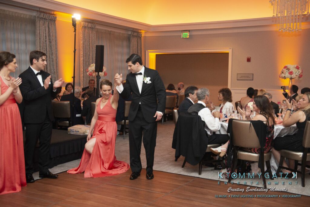 A man in a tuxedo escorts a woman in a red dress through a formal event room with seated guests clapping.