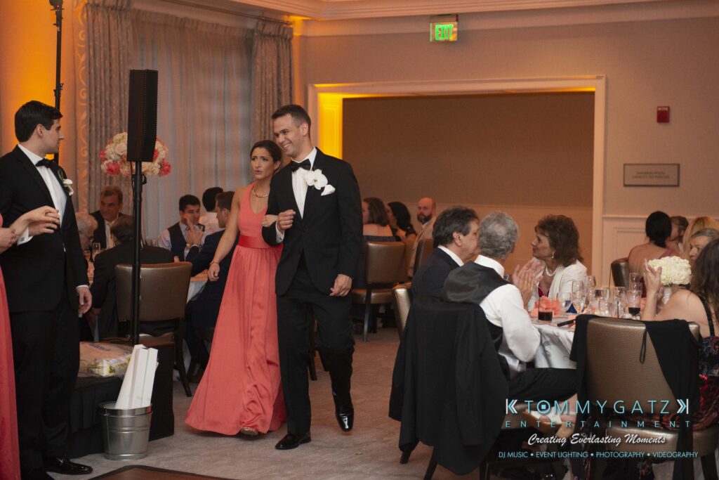 A couple in formal attire walks into a banquet room filled with seated guests. The room is decorated with flowers and soft lighting.