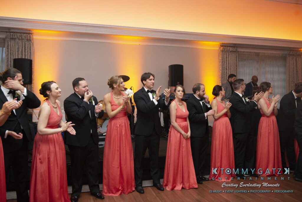 A group of people in formal attire, some in black suits and others in matching coral dresses, stand side by side in a room with orange lighting, clapping and facing forward.