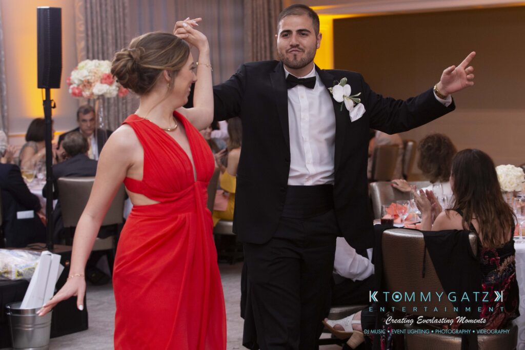 A man in a tuxedo and a woman in a red dress dance together in an indoor event setting.
