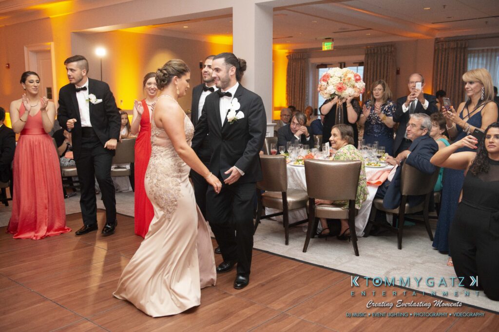 People dressed formally are dancing in a banquet hall while others sit and stand, watching and taking photos. Tables are set with floral centerpieces.