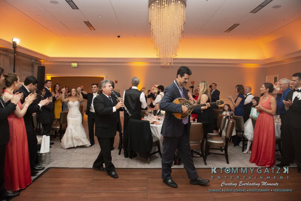 A well-dressed crowd claps as musicians play. A bride stands nearby, and guests at tables watch the performance under a large chandelier in a warmly lit room.