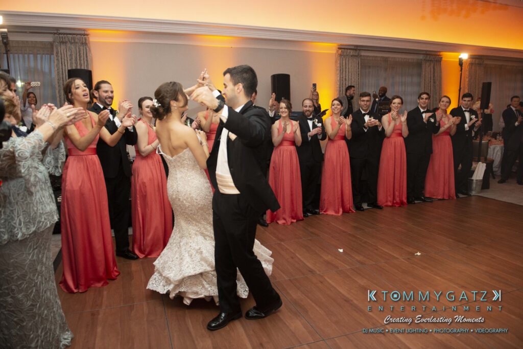 A bride and groom dance in a ballroom while guests in formal attire cheer and clap. A line of bridesmaids in coral dresses stands nearby.