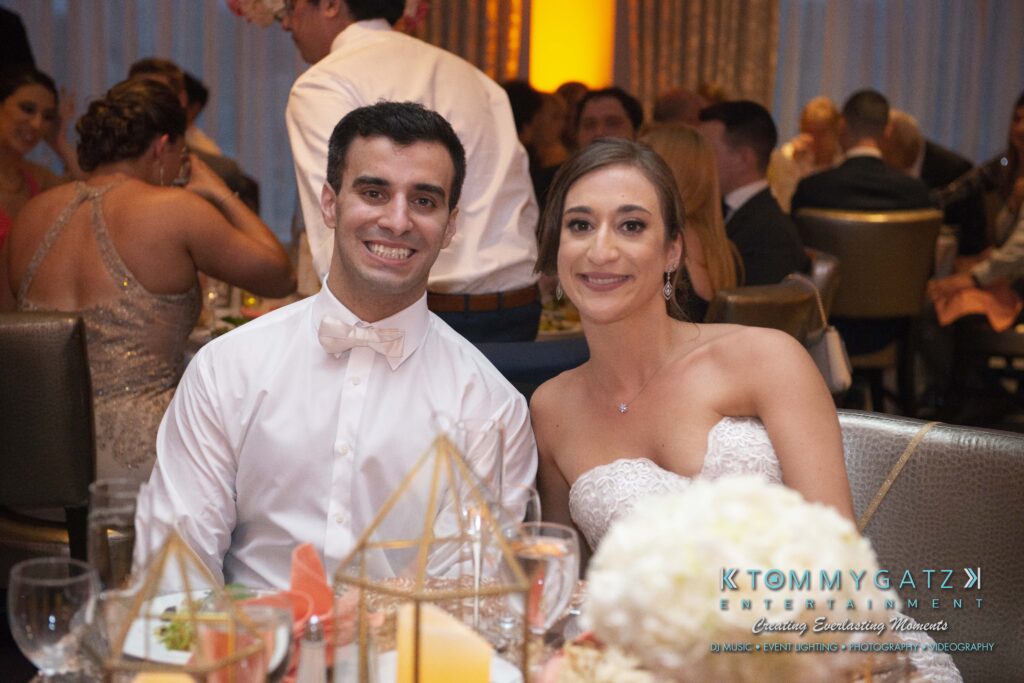 A couple, dressed formally, sits smiling at a table during a wedding reception. Guests are seated in the background.