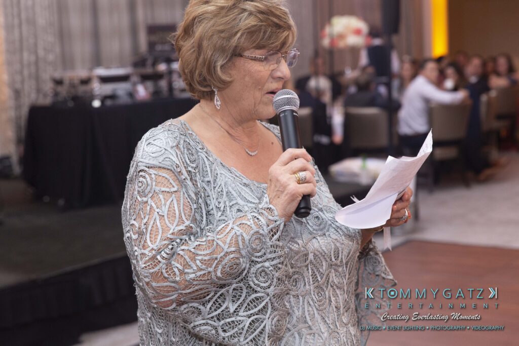 An older woman in a patterned dress speaks into a microphone while holding a piece of paper at an indoor event.