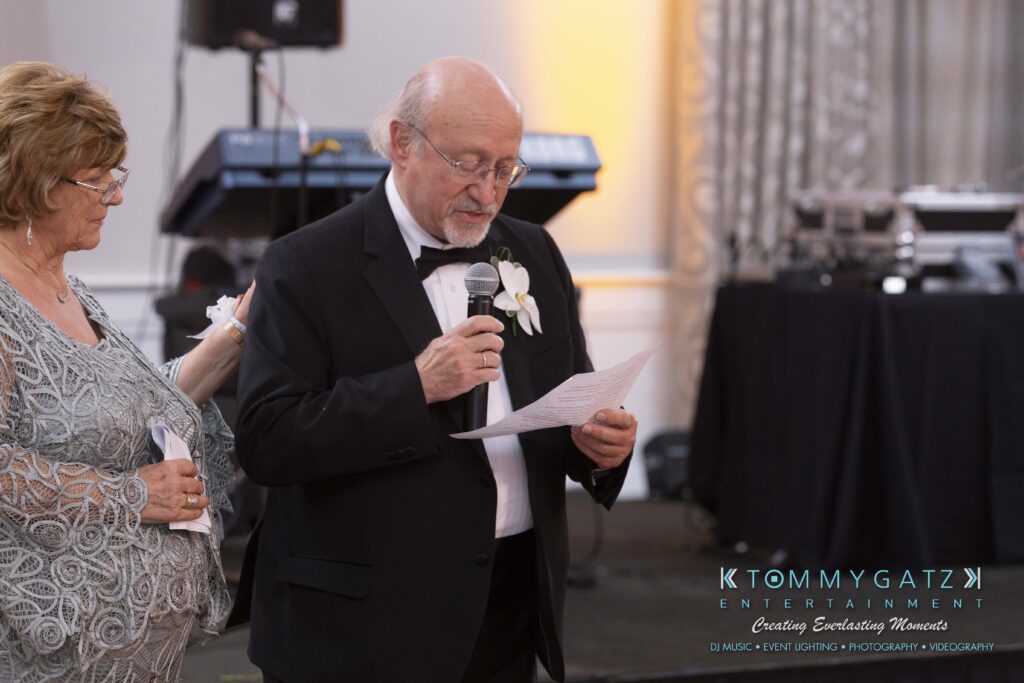 An elderly man in a black tuxedo reads a speech from a paper, standing beside a woman. They're in a decorated event space with musical equipment in the background.