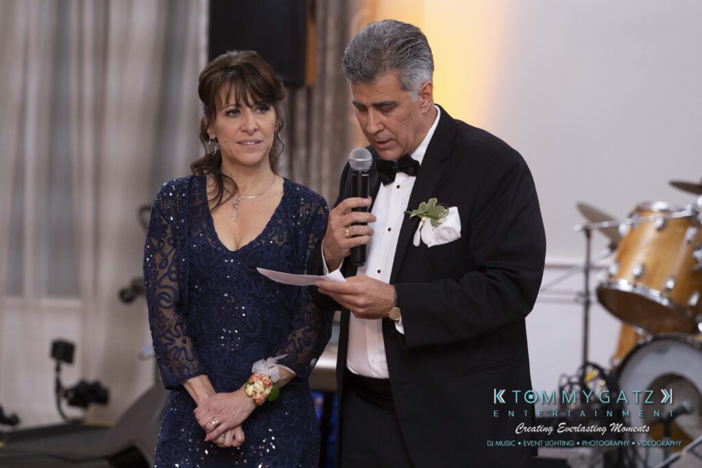 A woman and a man in formal attire stand together. The man reads from a paper while speaking into a microphone at an event. Musical instruments are in the background.