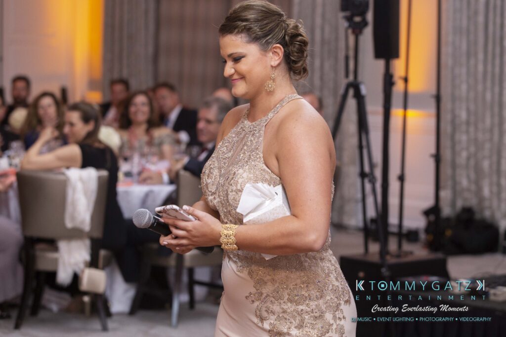 A woman in a formal dress holds a microphone and clutch, standing in a room with seated guests.