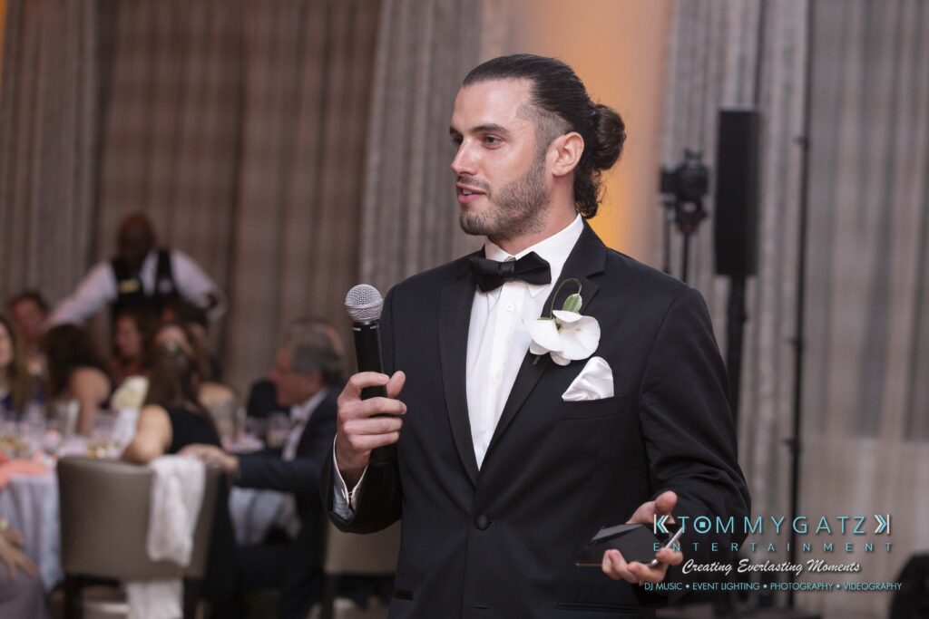 A man in a tuxedo speaks into a microphone at a formal indoor event. Guests are seated at tables in the background.