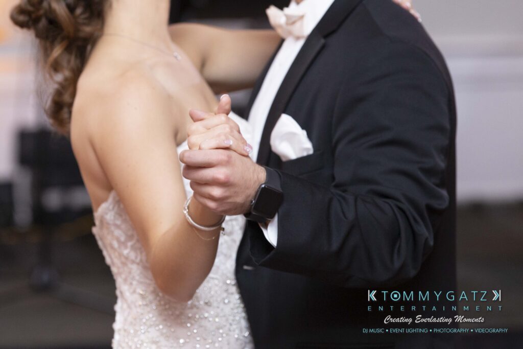 A couple dancing together, the woman in a strapless dress and the man in a suit. Their hands are clasped, with the man wearing a watch.